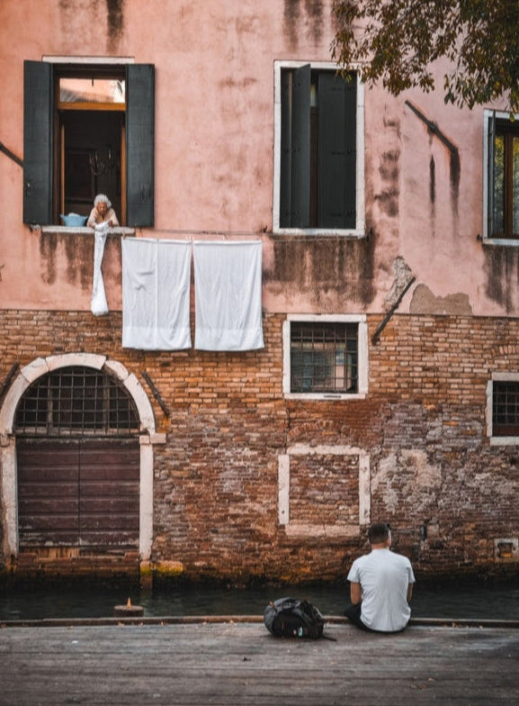 Two Strangers in Venice fine art photograph showing an elderly woman leaning from a Venetian window while a lone figure sits beside a quiet canal below.